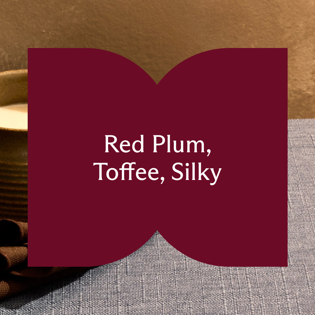 A close-up graphic overlay with a dark red background shaped like two joined arches. The white text reads: "Red Plum, Toffee, Silky". The background shows a portion of a blue textured tablecloth.