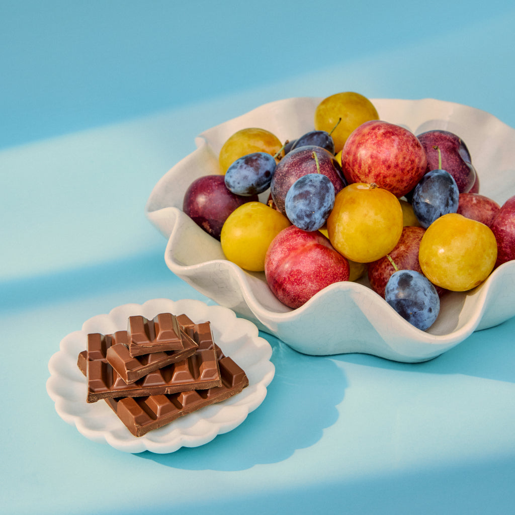 Bowl of plums with a small plate of chocolate bars on a blue background