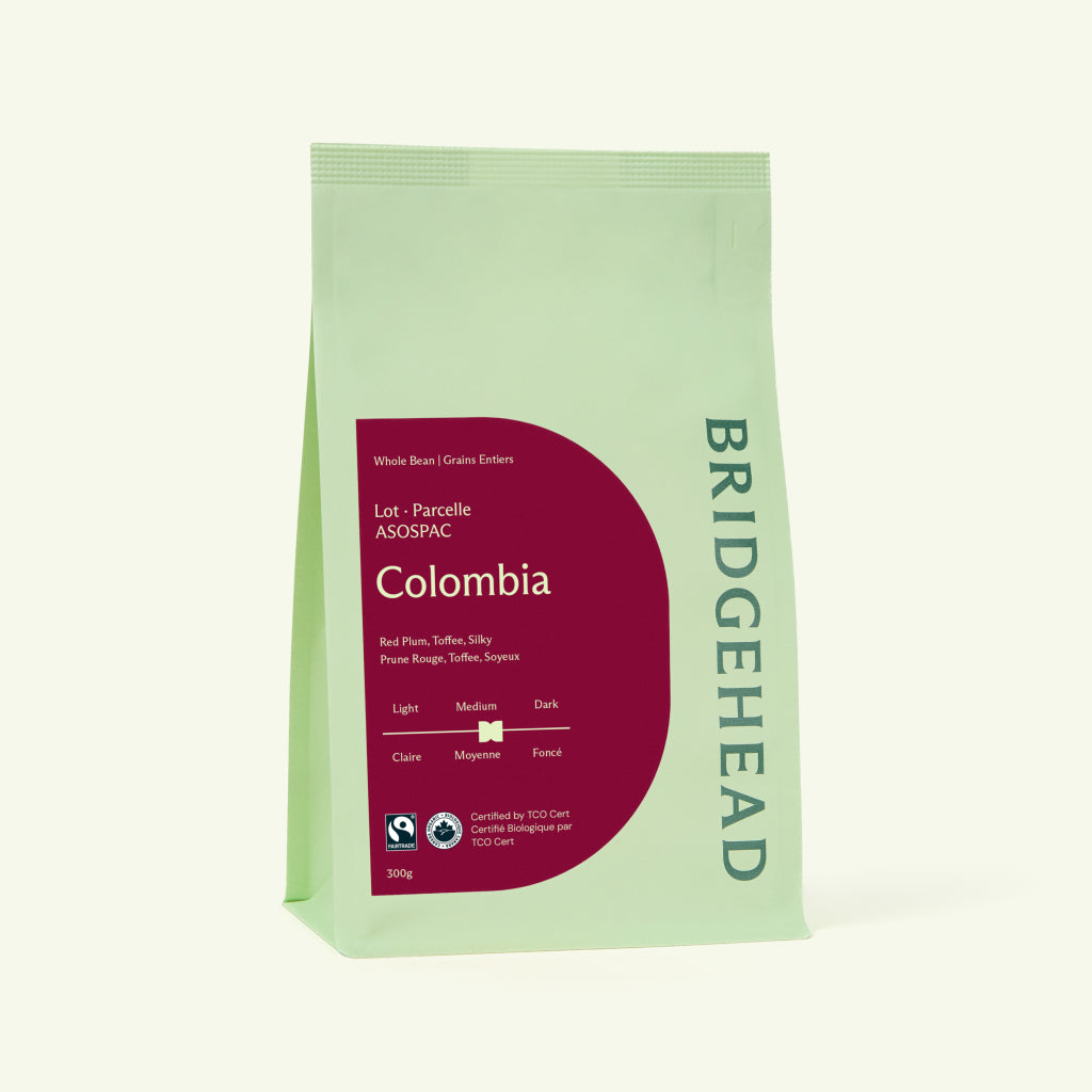 A product shot of a light green Bridgehead Colombia coffee bag. The bag features a deep magenta half-circle label indicating a Medium roast with tasting notes of red plum, toffee, and silky.