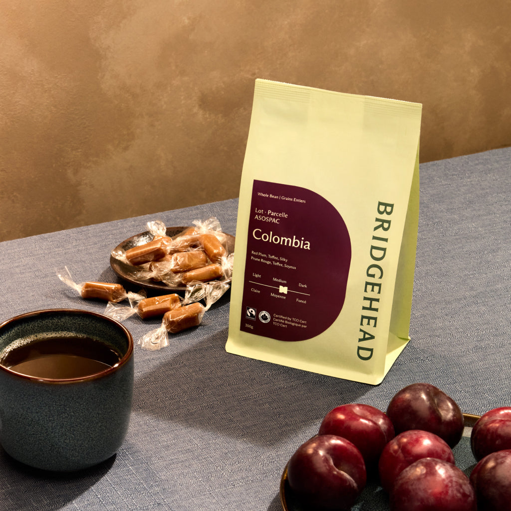 A lifestyle photograph featuring a bag of Bridgehead Colombia whole bean coffee with a deep purple label on a blue textured tablecloth. The scene includes a dark blue ceramic mug of coffee, a small plate of wrapped toffee candies, and several fresh red plums. The lighting is warm against a neutral, textured wall background.